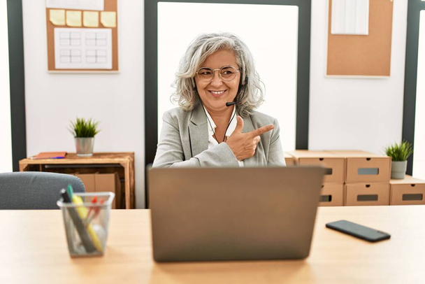 Bizneswoman w średnim wieku siedzi na biurku pracując za pomocą laptopa w biurze wesoły z uśmiechem twarzy wskazując z ręki i palca w górę do boku ze szczęśliwym i naturalnym wyrazem twarzy  - Zdjęcie, obraz