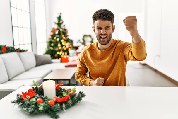 クリスマスツリーの怒りによってテーブルの上に座っているアラブの若い男と怒りを叫びながら拳をフラストレーションと激怒調達怒っている。怒りと攻撃的な考え方.  - 写真・画像