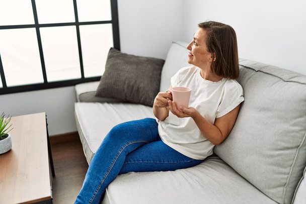 Hiszpanka w średnim wieku siedzi na kanapie i pije kawę w domu. - Zdjęcie, obraz