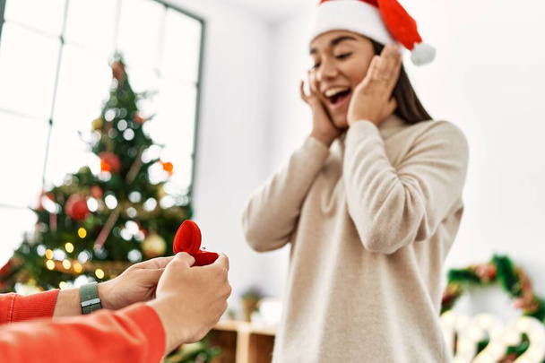 Νέοι ισπανόφωνοι γιορτάζουν τα Χριστούγεννα με πρόταση γάμου κρατώντας δαχτυλίδι αρραβώνων στο σπίτι. - Φωτογραφία, εικόνα