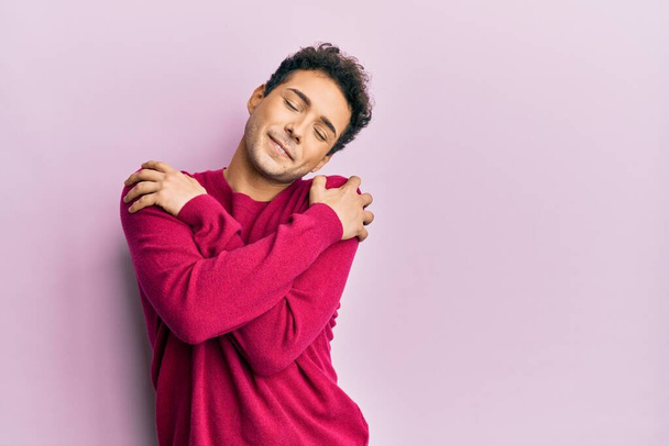 Handsome hispanic man wearing casual clothes over pink background hugging oneself happy and positive, smiling confident. self love and self care  - Fotoğraf, Görsel