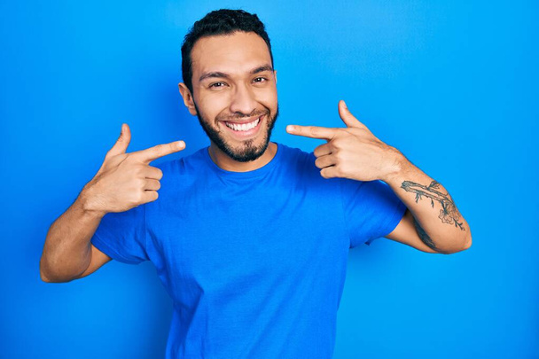 Homme hispanique à la barbe vêtu d'un t-shirt bleu décontracté souriant et joyeux montrant et pointant du doigt les dents et la bouche. concept de santé dentaire.  - Photo, image