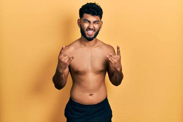 Arab man with beard wearing swimwear shirtless shouting with crazy expression doing rock symbol with hands up. music star. heavy concept.  - Photo, Image