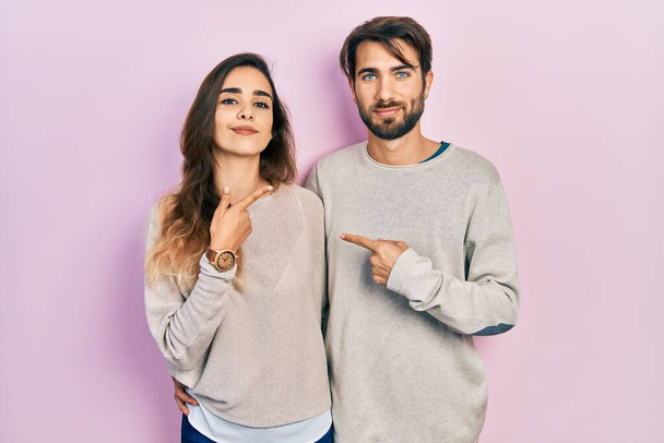 Joven pareja hispana vestida con ropa casual apuntando con el dedo de la mano hacia un lado mostrando publicidad, cara seria y tranquila  - Foto, imagen