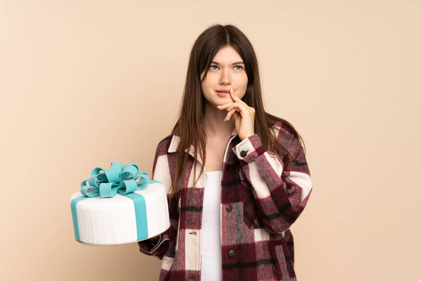 Young Ukrainian girl holding a big cake isolated on beige background having doubts and thinking - Фото, изображение