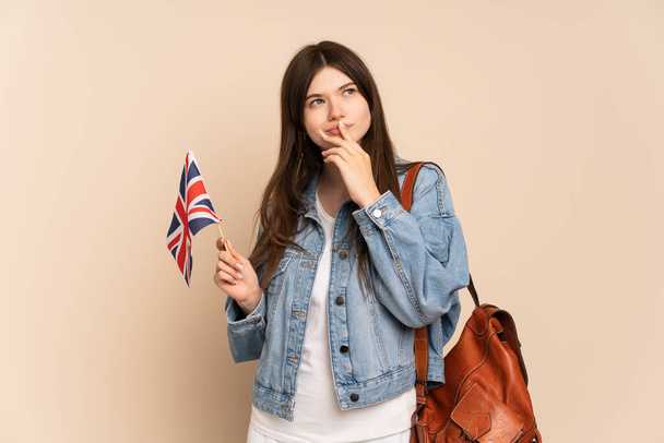 Young Ukrainian girl holding an United Kingdom flag isolated on beige background having doubts while looking up - Photo, Image