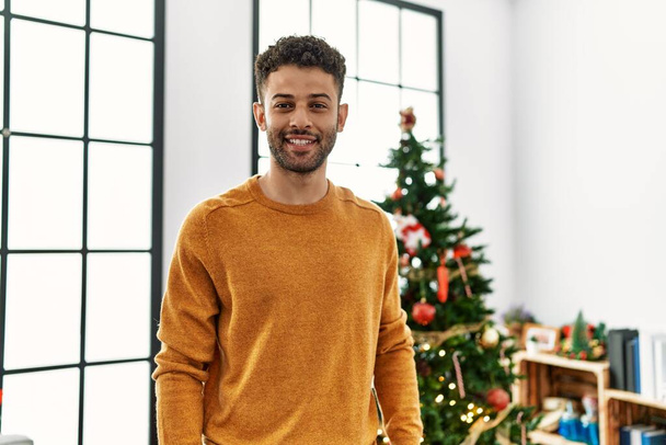 Young arab man smiling happy standing by christmas tree at home. - Photo, Image