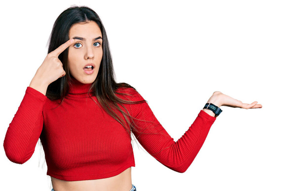 Giovane adolescente bruna indossa maglione rosso collo alto confuso e infastidito con palmo aperto che mostra spazio copia e punta il dito sulla fronte. Pensaci..  - Foto, immagini