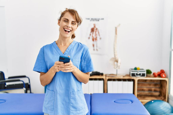 Joven fisioterapeuta caucásica chica sonriendo feliz usando smartphone en la clínica. - Foto, imagen