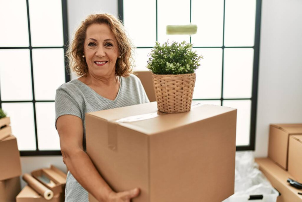Keski-ikäinen valkoihoinen nainen hymyilee onnellinen pitämällä pahvilaatikkoa uudessa kodissa. - Valokuva, kuva