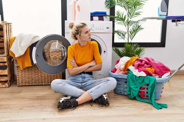 Jovem loira fazendo lavanderia sentado pela máquina de lavar roupa olhando para o lado com os braços cruzados convencido e confiante  - Foto, Imagem