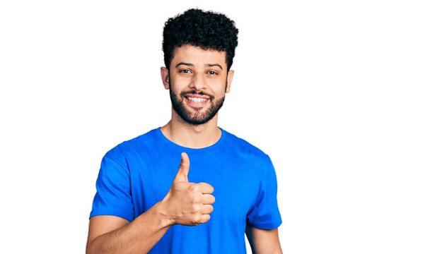 ひげを生やした若いアラブ人男性は、幸せな親指を手でジェスチャーするカジュアルな青のTシャツを着ています。成功を示すカメラを見ると.  - 写真・画像