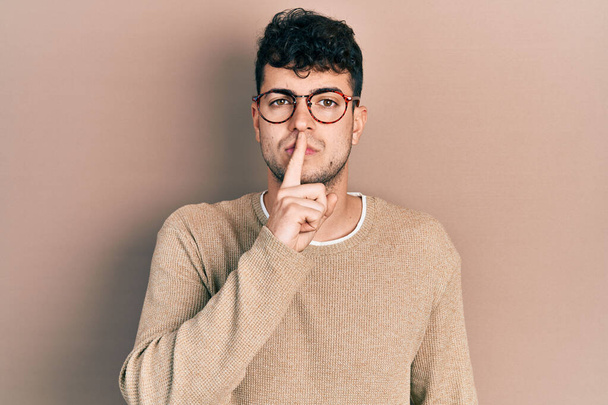 Νεαρός Ισπανός που φοράει καθημερινά ρούχα και γυαλιά και ζητάει ησυχία με το δάχτυλο στα χείλη. σιωπή και μυστική έννοια.  - Φωτογραφία, εικόνα