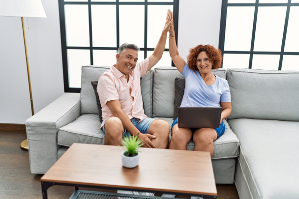 Középkorú férfi és nő pár segítségével laptop pacsi felemelt kézzel ül kanapén otthon - Fotó, kép