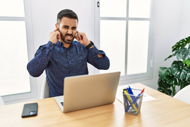 Jonge Spaanse man met baard die op kantoor werkt met laptop die oren bedekt met vingers met geïrriteerde expressie voor het lawaai van luide muziek. doof concept.  - Foto, afbeelding