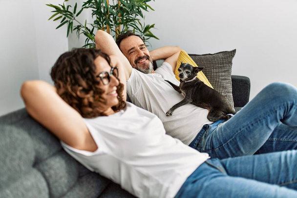 Hispánský pár středního věku, usmívající se šťastně, se uvolnil s rukama za hlavou. Sedět doma na gauči se psem. - Fotografie, Obrázek
