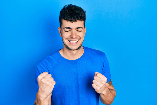 Nuori latinomies yllään rento sininen t paita erittäin onnellinen ja innoissaan tekee voittaja ele kädet nostetaan, hymyillen ja huutaen menestystä. juhla käsite.  - Valokuva, kuva
