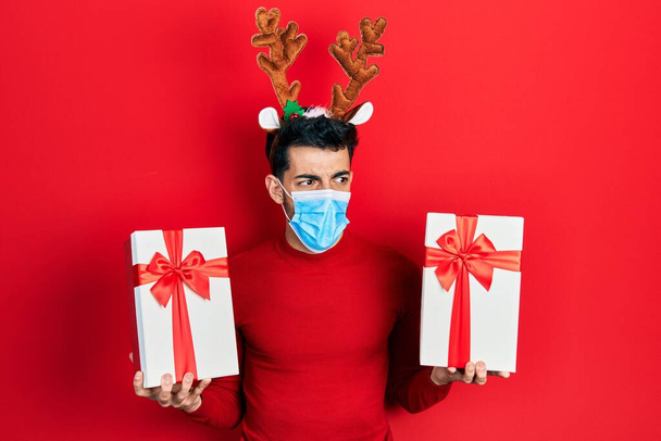 可愛いクリスマスのトナカイの角と医療用マスクを身に着けている若いヒスパニック系の男は怒りと怒りを叫び、失望と激怒を叫んで贈り物を保持しています。怒りと攻撃的な考え方.  - 写真・画像