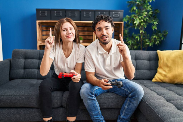 Młoda para grająca w kontrolera gier wideo w domu zaskoczona pomysłem lub pytaniem wskazującym palcem szczęśliwą twarz, numer jeden  - Zdjęcie, obraz
