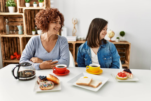 Family of mother and down syndrome daughter sitting at home eating breakfast looking away to side with smile on face, natural expression. laughing confident.  - Zdjęcie, obraz