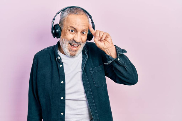 Handsome senior man with beard listening to music using headphones pointing finger up with successful idea. exited and happy. number one.  - Foto, afbeelding