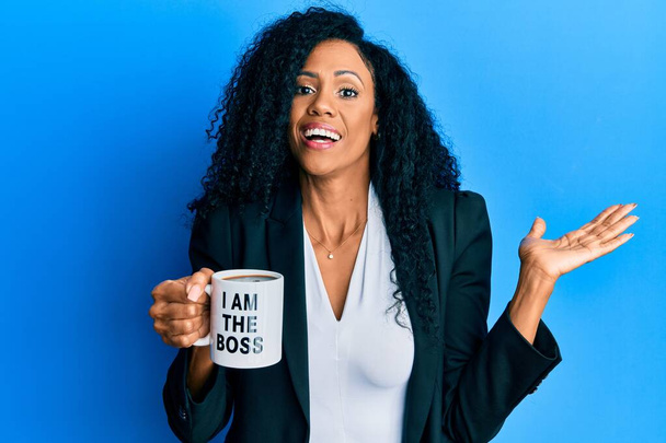Orta yaşlı Afro-Amerikalı kadın ben patron kahve fincanından içiyorum başarıyı mutlu bir gülümsemeyle ve kazanan ifadeyle el kaldırarak kutluyorum.  - Fotoğraf, Görsel