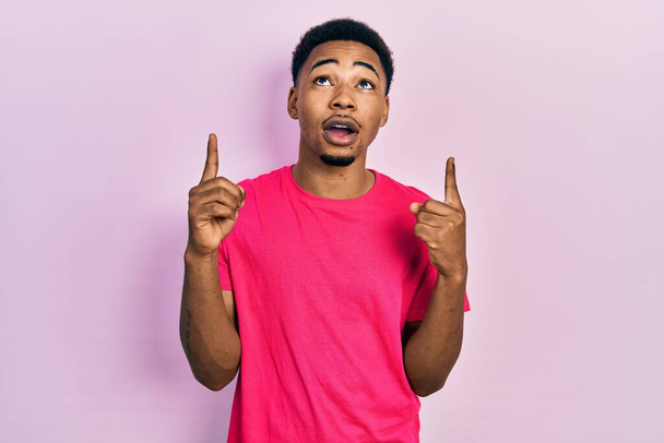Молодой африканский американец в футболке с надписью casua удивлен и удивлен, глядя вверх и указывая пальцами и подняв руки.  - Фото, изображение