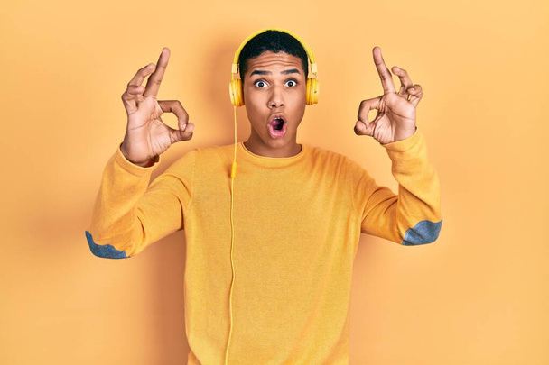 Young african american guy listening to music using headphones looking surprised and shocked doing ok approval symbol with fingers. crazy expression  - Fotó, kép