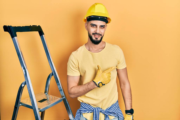 Όμορφος άντρας με γενειάδα από κατασκευαστικές σκάλες φορώντας καπέλο χαρούμενο με χαμόγελο στο πρόσωπο δείχνοντας με το χέρι και το δάχτυλο στο πλάι με χαρούμενη και φυσική έκφραση  - Φωτογραφία, εικόνα