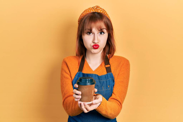 Zrzka mladá žena v zástěře servírky drží odnést šálek kávy foukání tváře s legrační tvář. ústa nadýchaná vzduchem, lapající vzduch.  - Fotografie, Obrázek
