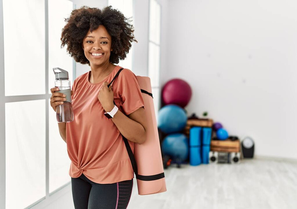 Young african american woman smiling confident holding yoga mat and bottle of water at sport center - Zdjęcie, obraz