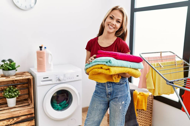 Mulher caucasiana jovem segurando lavandaria limpa olhando positivo e feliz de pé e sorrindo com um sorriso confiante mostrando dentes  - Foto, Imagem
