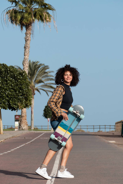 Junge Skaterin mit Afrohaarfrisur hält Skateboard, lächelt, blickt in die Kamera - Foto, Bild