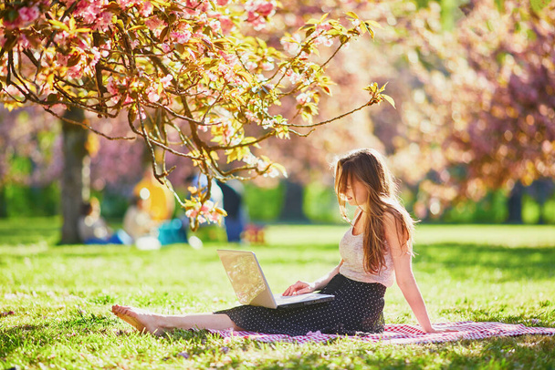 Beautiful young girl working on her laptop in park during cherry blossom season. Young woman in famous Park of Sceaux near Paris, France. Freelance, distance learning or remote work concept - Fotó, kép
