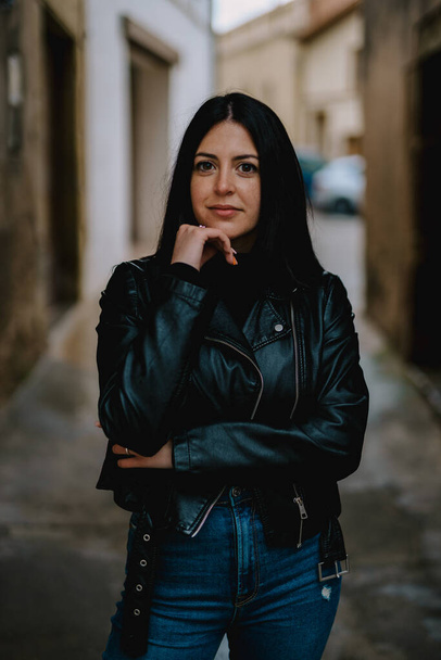 portrait of a beautiful young woman in a black leather jacket and jeans posing on the street - Foto, imagen