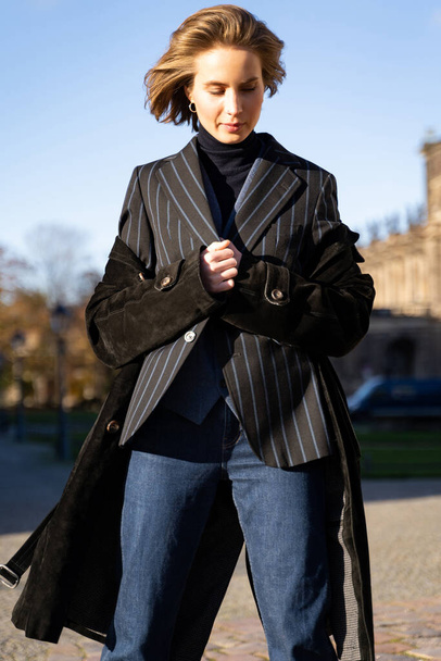 Young blond short-haired woman in striped jacket, jeans and coat on sunny street - Photo, image