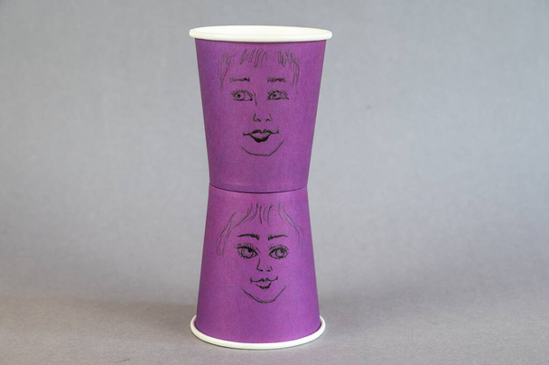 Два фиолетовых стакана с нарисованными лицами. Мужские и женские лица на одноразовых стаканах горячих напитков. Крупный план. - Фото, изображение