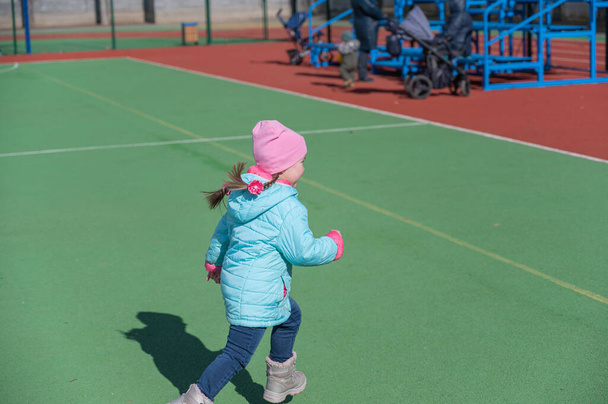 Ребенок в синей куртке бегает по зеленой спортивной площадке. Родители и бабушка с детскими колясками на заднем плане. Злобная пятилетняя девочка с косичками резвится. Детство - Фото, изображение
