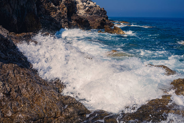 Wonderful views of the blue Mediterranean Sea. Sunny rocks, waves with foam and splashing water. The wave crashes into the rocks on the shore - Fotografie, Obrázek