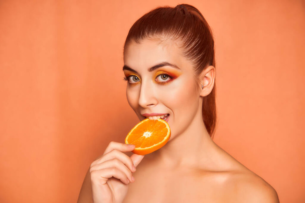 Portrait of beautiful young woman biting an orange on yellow background.  - Photo, Image