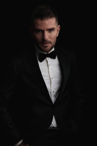 portrait of confident businessman in black tuxedo holding hands in pockets on black background - Foto, immagini