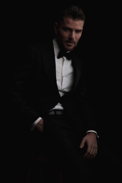 portrait of sexy best man in tuxedo holding hand in pocket, being confident while sitting in studio in front of black background - Zdjęcie, obraz