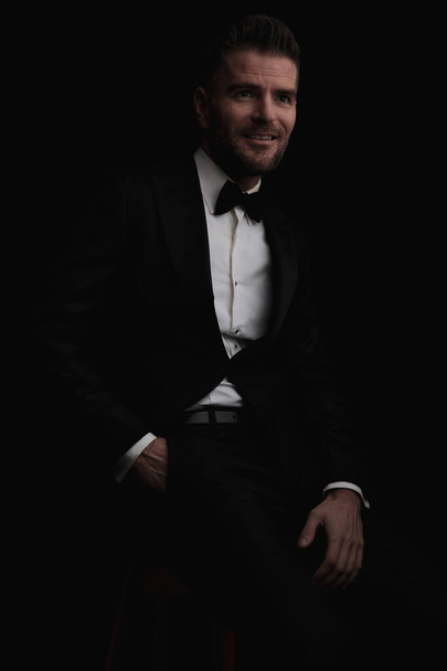 happy groom with hand in pocket wearing black elegant tuxedo and looking away for his bride while sitting on black background - Photo, Image
