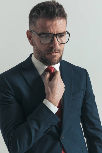 portret van ongeschoren zakenman in de veertig met bril weg te kijken terwijl de vaststelling van rode stropdas in de voorkant van grijze achtergrond in de studio - Foto, afbeelding