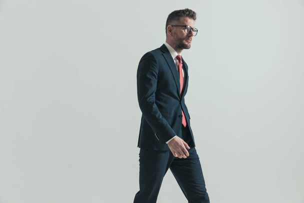 side view of middle aged man with grizzled hair and glasses looking to side and walking in front of grey background in studio - Photo, Image