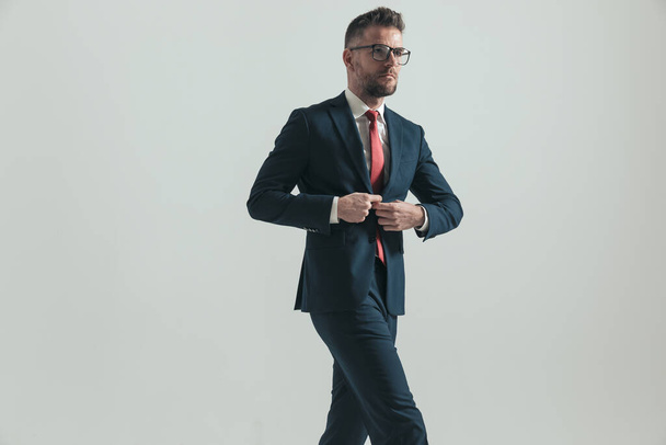 selbstbewusster bärtiger Mann mit Brille knöpft eleganten Anzug an, während er isoliert auf grauem Hintergrund im Studio wandelt - Foto, Bild