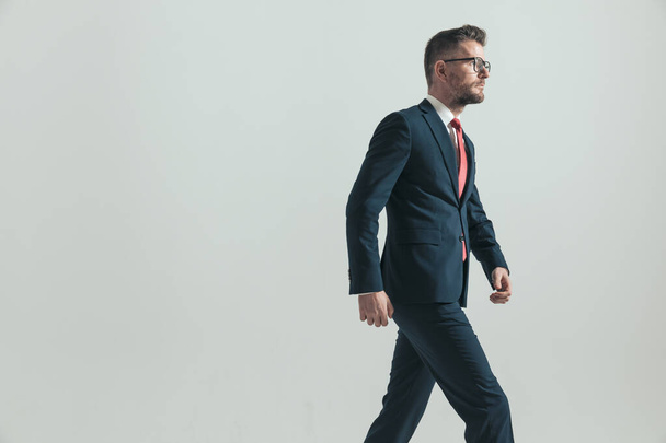side view of elegant middle aged man in suit with glasses walking and looking to side on grey background - Foto, imagen