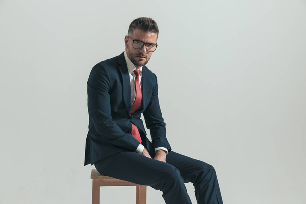 sexy man with eyeglasses and elegant suit sitting on wooden chair in a side view pose on grey background - Фото, изображение