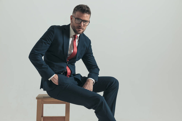 vue latérale d'un homme d'affaires assis avec des lunettes tenant la main dans la poche et posant devant un fond gris  - Photo, image