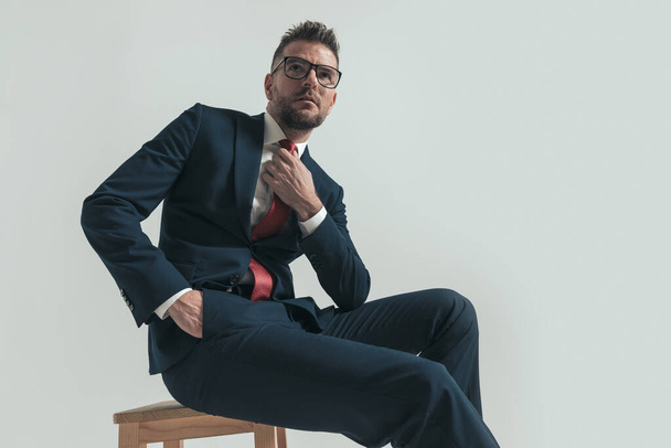 όμορφος επιχειρηματίας με γυαλιά με κοστούμι κοιτάζει ψηλά, προσαρμόζοντας τη γραβάτα και κρατώντας το χέρι στην τσέπη, ενώ κάθεται σε ξύλινη καρέκλα στο στούντιο - Φωτογραφία, εικόνα